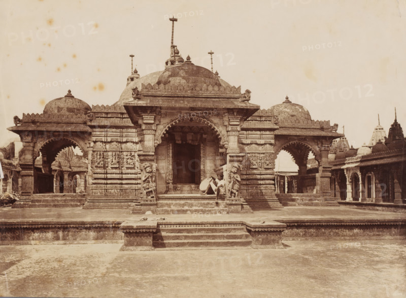Temple complex, Palitana, Gujarat, India, photograph, Palitána ?, India., inscription: 'Palitana', c. 1870 - c. 1900, photographer, paper, albumen print, height 239 mm x width 177 mm, A wide stone temple facade with domed roofs and carved pillars sits centered on a raised platform accessed by a short flight of steps, flanked by arched porticoes on either side and smaller domed structures in the background; ornate sculptural reliefs decorate the walls and pillars, and two carved guardian figures stand beside the central entrance where a seated person rests against one pillar holding an object; the foreground shows a broad paved courtyard leading to the temple and scattered small objects on the steps and platform.