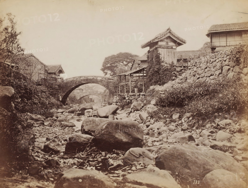 The Amidabashi Bridge and behind it the Koraibashi Bridge over the Nakashimagawa River, Nagasaki, Japan, photograph, The photo was taken downstream. Nearby was the house of Ueno Hikoma. See Nagasaki University Library Collection, where the same photo (colored)., number: '388', inscription: 'illegible', c. 1870 - c. 1900, photographer, paper, albumen print, height 195 mm x width 258 mm, Kyushu, A sepia-toned scene shows a rocky streambed extending toward the background with smooth boulders and smaller stones scattered along the water and banks, wooden tubs and buckets resting among the rocks, a stone arched bridge crossing the stream with a second distant arch visible beyond it, timber and stone buildings with tiled roofs lining the banks including a tall wooden structure partially covered in climbing vegetation, a low wooden shelter with posts near the water, a stone retaining wall supporting a slope on one side, patches of grass and shrubs on the banks, and a large tree with a broad c...