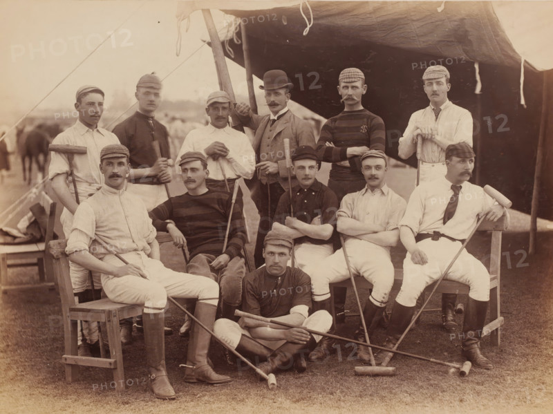 Polo players, Abbottabad, Khyber Pakhtunkhwa, Pakistan, album page, photograph, Group portrait of polo players, Pakistan. The men are sitting and standing partly under a tarpaulin., number: '1560', title: 'Polo Players Abbottabad 1885', 1885, photographer, pasteboard, paper, albumen print, height 207 mm x width 281 mm, height 264 mm x width 347 mm, A group of men posed in front of a tent, wearing a mix of light and dark shirts, jackets, and caps, many with mustaches and short hair, some seated on wooden chairs or benches and others standing behind; several men hold long wooden mallets and sticks across their laps or resting on the ground, while one man in a bowler hat stands with an arm raised on a pole supporting the tent; the tent fabric hangs overhead with visible ropes and ties, and the grassy ground beneath shows scattered mallet heads and the lower legs of the seated men in tall boots.