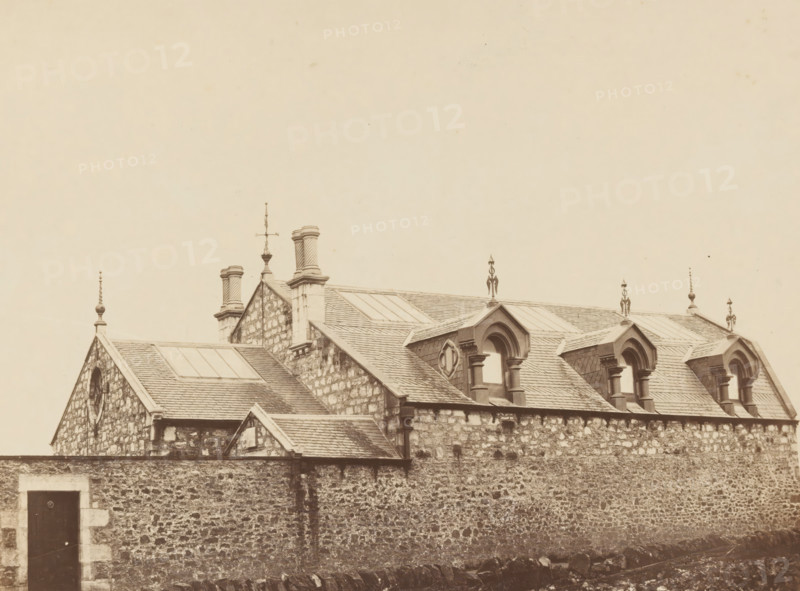 View of country house in England, photograph, England, c. 1875 - c. 1885, photographer, pasteboard, paper, albumen print, albumendruk, height 196 mm x width 262 mm, height 402 mm x width 582 mm, farm or solitary house in landscape, A sepia-toned view of a stone building with a tiled roof and multiple gables, featuring decorative roof finials and chimney stacks, dormer windows with arched tops and columns, rectangular skylights set into the roof, and a high rough stone wall in the foreground with a dark wooden door set into a lighter stone frame.