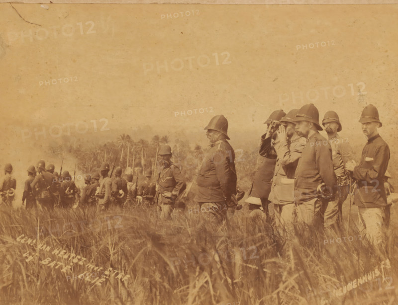 General van Heutsz with staff in Aceh during the attack on the benteng complex Batoe Iliq in the coastal state of Samalanga, photograph, General van Heutsz with staff in Aceh., inscription: 'Gen. van Heutsz with staff, at Baté Illië, C. Nieuwenhuis ft.', photographer: Christiaan Benjamin Nieuwenhuis, Aceh, 1901-03-12, Sumatra, pasteboard, paper, albumen print, albumendruk, height 218 mm x width 284 mm, height 326 mm x width 440 mm, Person, Johannes Benedictus van Heutsz, commander-in-chief, general, marshal, binoculars, spy-glass, A sepia-toned photograph showing a group of men in military uniforms and pith helmets standing and facing toward the left, some holding rifles and one using binoculars, with tall grasses in the foreground and a line of palm trees visible in the middle background.