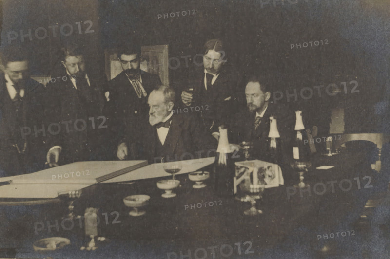 George Hendrik Breitner and others, looking at a portfolio, possibly during an art viewing at Arti et Amicitiae, photograph, Amsterdam, 1900 - 1923, photographer, paper, pasteboard, baryta paper, gelatin silver print, developing gelatin silver print, height 153 mm x width 227 mm, Person, George Hendrik Breitner, historical persons, painter in non-work situation (circle, club of artists), Arti et Amicitiae, A dim, sepia-toned scene of men gathered around a table with documents and tableware; several men in dark suits and bow ties stand and sit closely together, one older man with a white beard seated near the center leaning over a large sheet on the table while others look on, some holding small glasses; the table foreground shows bowls, goblets, bottles with paper coverings, a folded cloth with an image, and scattered small dishes and glassware, with a chair partially visible at the right and a framed object on the wall behind the group.