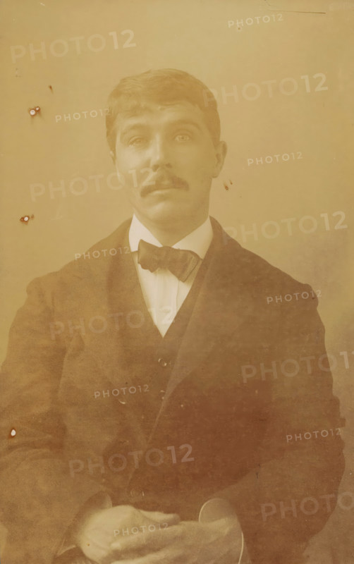 Portrait of the artist Rik Wouters, photograph, 1915-10, photographer, paper, baryta paper, daylight gelatin silver print, height 170 mm x width 106 mm, Person, Rik Wouters, historical persons, portrait, self-portrait of painter, A sepia-toned portrait of a man shown from the waist up wearing a dark suit jacket over a vest and a white shirt with a bow tie, his hair neatly combed and a mustache on his upper lip, his hands resting in his lap with one hand holding a hat, and small blemishes and spots visible on the photograph surface.