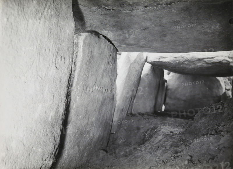 Hunebed van Kerguntuil, Trégastel, photograph, inscription: 'IX, Ban v Heuvel, Allée Couverte de Kerguntuil, Trégastel C. du N., sculpture on two supporting stones', photographer: attributed to Jan Lanting, supervision: Albert Egges van Giffen, 1939-05-27 - 1939-06-04, Attributed to, paper, baryta paper, gelatin silver print, developing gelatin silver print, height 123 mm x width 172 mm, megalithic chambered tomb, dolmen, A close-up interior view of a narrow stone passage showing rough, weathered vertical slabs forming the sides and flat horizontal slabs forming the ceiling, with visible stone textures, joints, and chipped edges; the ground is uneven and covered in compacted dirt and small debris; light enters from an opening at the far end, illuminating the irregular surfaces and casting soft shadows along the stones.