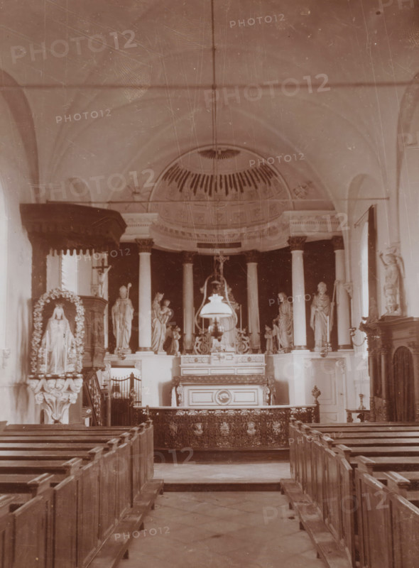 Interior of the St. Lambertus Church in Huisseling, photograph, annotation: 'Municipality of Ravenstein, Roman Catholic church in Huiselinge (NB) near Ravenstein March 1911', Huisseling, 1911, photographer, Attributed to, paper, pasteboard, daylight collodion silver print, height 218 mm x width 161 mm, interior of church, A sepia-toned interior view of a church nave with wooden pews leading toward an ornate altar area, flanked by tall columns and statues on raised pedestals; a domed ceiling with decorative molding is visible above the altar, and a hanging lamp or chandelier is suspended in front of the altar; carved woodwork, decorative panels, and sculptural figures, including a draped statue in a floral arch on one side, are arranged symmetrically around the central altar; railings and a patterned altar frontal occupy the foreground, and high arched walls and windows frame the scene.