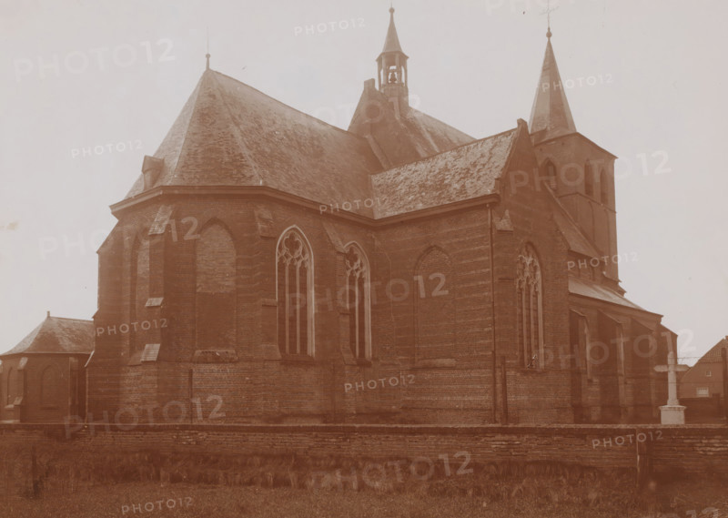 Northeast side of the St. Lambertus Church in Rosmalen, photograph, annotation: 'Rosmalen, March 1910', Rosmalen, 1910, photographer, Attributed to, paper, pasteboard, daylight collodion silver print, height 158 mm x width 221 mm, church (exterior), A large brick church building fills the image, showing steep pitched roofs covered in shingles, pointed spires, a small open bell tower, and tall narrow arched windows with tracery. The exterior walls display buttresses and recessed arched panels, and a low brick wall runs along the foreground with grass and vegetation in front of it. The overall tone of the photograph is sepia, and the sky appears pale and empty above the building.
