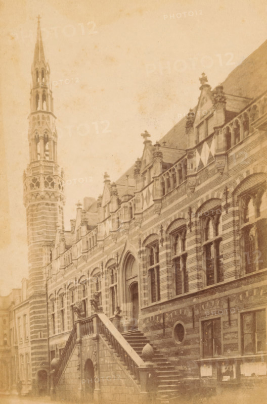 Hotel de Ville d'Alkmaar, Exterior of the Alkmaar Town Hall, photograph, Alkmaar, c. 1865 - c. 1880, photographer, paper, pasteboard, albumen print, height 147 mm x width 97 mm, town hall, façade (of house or building), Town Hall (Alkmaar), A sepia-toned photograph of a large ornate stone building with striped masonry and arched windows, featuring an exterior staircase with stone balustrades leading up to an arched entrance flanked by sculpted figures; the façade shows decorative gables and window tracery, triangular flags hanging from the upper level, and sculpted finials along the roofline; a tall, intricately carved tower with openwork sections rises beside the building, and rows of windows and masonry details extend along the street-level façade.