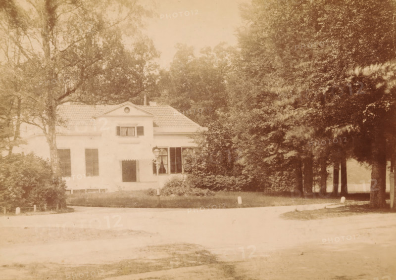 Exterior of a country house on the Zwolseweg in Apeldoorn, photograph, inscription: 'Ant. C. Reelick Photographer Apeldoorn. Special facility for enlargements of any portrait.', photographer: AC Reelick, Apeldoorn, c. 1880 - c. 1900, paper, pasteboard, albumen print, height 97 mm x width 138 mm, rural housing, eg country-house, villa, cottage, façade (of house or building), A two-story light-colored house with a tiled roof is partially obscured by trees and shrubs; the front facade shows shuttered windows and a small central dormer with an oval opening, a doorway with an awning, and a lamp mounted near a ground-level window. Tall leafy trees surround the building, with a grove of darker-trunked trees to the right forming a shaded area, and low hedges and bushes in front. A paved road curves across the foreground with small posts along its edge, and the scene has a soft, faded sepia tone.