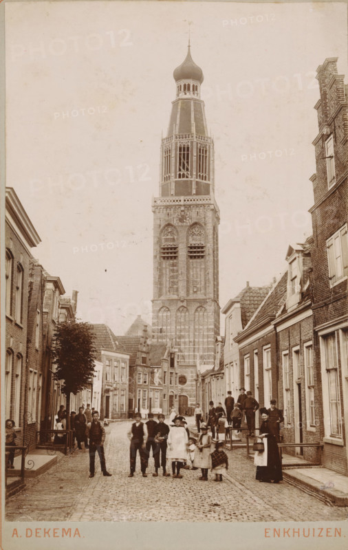 View of Torenstraat in Enkhuizen, with the Zuiderkerk in the background, photograph, inscription: 'Enkhuizen', photographer: Albert Dekema, Enkhuizen, c. 1900, paper, pasteboard, daylight gelatin silver print, height 148 mm x width 97 mm, street, church (exterior), Zuiderkerk (Enkhuizen), A narrow cobblestone street lined with brick buildings on both sides leads toward a tall ornate tower with arched openings and a bulb-shaped dome at the top, dominating the center background. People of various ages stand and walk along the street, including groups of children and adults clustered near the foreground and along the sidewalks, some wearing hats and long coats and a woman holding a basket. Windows and doorways of the adjacent buildings are visible, as are rooftops and a tree on the left side. The scene is captured in a sepia-toned photograph with a clear vertical composition emphasizing the tower.