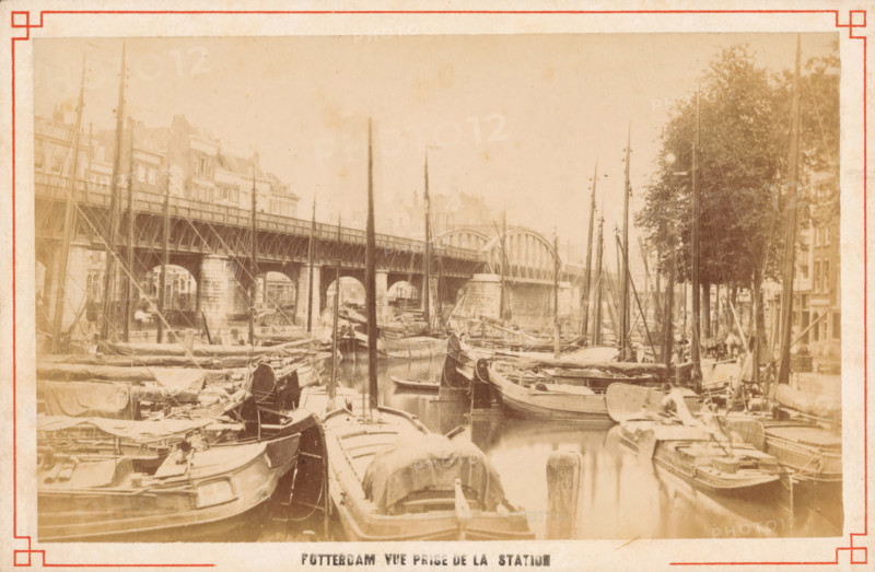 Boats on the Kolk in Rotterdam, Rotterdam vue prize de la station, photograph, Rotterdam, c. 1880 - c. 1900, photographer, paper, pasteboard, albumen print, height 63 mm x width 149 mm, canals, waters (in city), ships (in general), Kolk, A sepia-toned waterfront scene showing numerous moored wooden boats with tall masts filling a narrow canal, a metal arched bridge spanning the water in the background, stone bridge supports and an elevated platform along the left side, multi-story buildings behind the bridge, a row of trees and additional buildings on the right bank, rigging and tarpaulins on many of the boats, and reflections of the boats and masts visible in the water.
