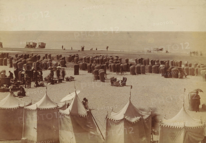Wicker beach chairs on the beach at Scheveningen, photograph, inscription: 'CP Wollrabe sen, The plate will be kept until reordered.', photographer: Carl Philip Wollrabe, Scheveningen, c. 1870 - c. 1885, paper, pasteboard, albumen print, height 101 mm x width 145 mm, beach (as place of recreation), chair, A wide sandy beach scene with rows of tall, rounded wicker beach chairs scattered across the shore, clusters of small tents with pointed tops and scalloped edges in the foreground, a few figures standing or walking near the waterline, some people seated in and beside the wicker chairs, a pair of wagon-like structures on the wet sand near the water, and a calm sea forming a horizontal backdrop.
