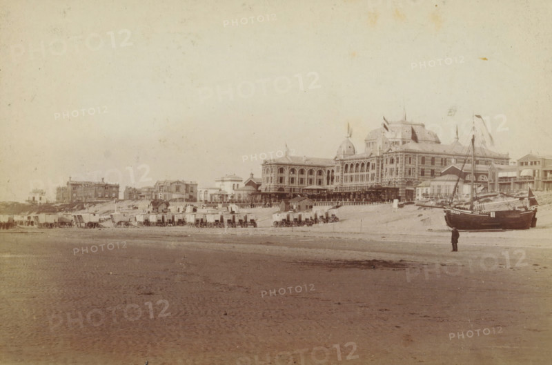 View of the beach at Scheveningen, with the Kurhaus on the right, photograph, inscription: 'CP Wollrabe sen., The plate is kept until reordered.', photographer: Carl Philip Wollrabe, Scheveningen, after 1887 - c. 1900, paper, pasteboard, albumen print, height 97 mm x width 145 mm, beach (as place of recreation), hotel, hostelry, inn, façade (of house or building), Kurhaus (Scheveningen), A wide sandy beach stretches across the foreground with rippled wet sand and scattered darker patches, leading up to a row of small wooden beach huts and horse-drawn caravans lined along the shoreline, each on wheels and clustered near the base of a low sandbank. Behind the huts a large ornate multi-story building with domed towers, arched windows, decorative facades and flags flying from its roof dominates the scene, flanked by several smaller classical and Victorian-style buildings and colonnades. Near the right side of the image a dark-hulled sailing boat with masts and rigging rests on the sand, an...