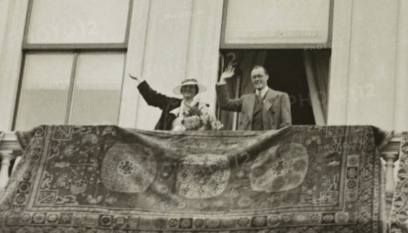 Engagement of Juliana, Queen of the Netherlands, and Bernhard of Lippe-Biesterfeld on the balcony of Noordeinde Palace in The Hague on September 8, 1936, photograph, Netherlands, 1936-09-08, photographer, paper, photography, height 88 mm x width 140 mm, The title mentioned here refers to the highest title obtained by the person portrayed, not necessarily to the title the person portrayed bears in this image., Person, Juliana of the Netherlands, Bernhard (Prince of the Netherlands), The Hague, Noordeinde Palace, Period, A grainy black and white photograph showing a man and a woman standing behind a balcony railing draped with an ornate patterned rug; the woman wears a wide-brimmed hat and a dark coat and holds a bouquet, while the man stands beside her in a suit and tie, both raising one hand in a waving gesture; behind them are tall window frames and curtains, and the rug in the foreground features large circular medallions and geometric border motifs.