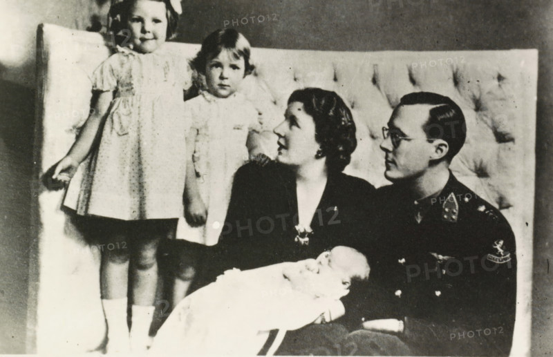Portrait of Juliana, Queen of the Netherlands, Bernhard van Lippe-Biesterfeld, Beatrix, Irene, Princess of the Netherlands, and Margriet, photograph, Bernhard, Irene and Margriet in Ottawa., Netherlands, 1943 - 1944, photographer, paper, photography, height 89 mm x width 132 mm, The title mentioned here refers to the highest title obtained by the person portrayed, not necessarily to the title the person portrayed bears in this representation., Person, Juliana of the Netherlands, Margriet (Princess of the Netherlands), North America, Ottawa, A black and white photograph shows a seated man in a dark uniform holding a swaddled infant, a seated woman beside him looking toward the man, and two young girls standing behind them on a cushioned backdrop. The standing girl on the left wears a light dress with short sleeves and a bow in her hair, while the other girl wears a lighter dress with a high collar and has short hair. The seated woman has short wavy hair and is wearing a dark outfit with...