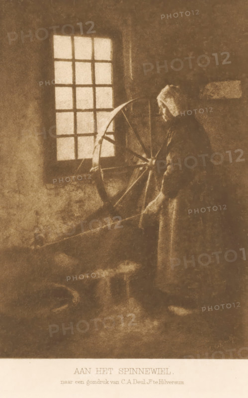 Woman by a spinning wheel, At the spinning wheel, page, photomechanical print, inscription: 'after a gum print by CA Deul Jr. in Hilversum.', photographer: Cornelis Albertus Deul, Netherlands, clichémaker: anonymous, c. 1900 - in or before 1905, paper, photoaquatint, height 167 mm x width 118 mm, Title page with subtitle is actually missing: subtitle will be the same for first, second, third and fifth series, farmers - AA - female farmer, farmer's wife, spinning-wheel, female sex, woman, A sepia-toned scene shows a person standing beside a large wooden spinning wheel near a window with multiple panes; the person wears a hooded garment and holds a distaff with both hands while gazing toward the wheel, and soft light from the window casts subtle shadows across a textured interior wall and floor.
