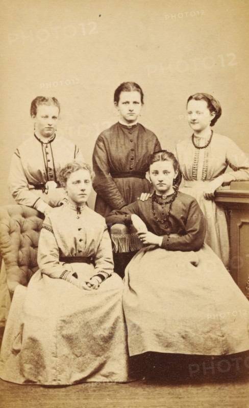 Portrait of a group of young women, one of whom is presumably the mother of AF Marmelstein, photograph, carte-de-visite, annotation: 'Grandma with friends, from Deventer', number: '1869.', photographer: Albertus Josephus van Poorten, Deventer, 1865 - 1869, paper, pasteboard, albumen print, albumendruk, height 101 mm x width 62 mm, anonymous historical persons portrayed in a group, in a group-portrait - BB - woman, Five young women are posed together in a sepia-toned studio photograph. Two women sit in the front on upholstered seats, wearing long skirts and high-collared bodices, one with her hands folded in her lap and the other resting an arm on the chair with a braided necklace and a pendant at her throat. Three women stand behind them, each wearing long dresses with fitted waists and buttoned bodices; the central standing woman has a darker dress and a direct gaze, while the two flanking her wear lighter dresses with decorative trim. An upholstered armchair is visible on the left an...