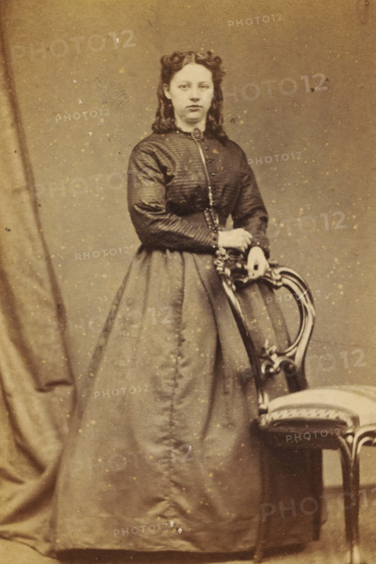 Portrait of an unknown young woman from the Marmelstein family, photograph, carte-de-visite, address: 'Photographisch Etablissement., S. van Caspel & Stapert., Bloemgracht DD 43., Amsterdam', photographer: Caspel & Stapert, S. van, Amsterdam, 1867 - 1870, paper, pasteboard, albumen print, albumendruk, height 100 mm x width 65 mm, historical persons not known by name - BB - woman, adolescent, young woman, maiden, A sepia-toned portrait of a young woman standing and resting her hands on the back of an ornate wooden chair with a cushioned seat, wearing a high-collared dress with a fitted, textured bodice and a full skirt, her hair styled in waves with coils at the top, a curtain draped to one side, and small spots and blemishes visible across the photograph surface.