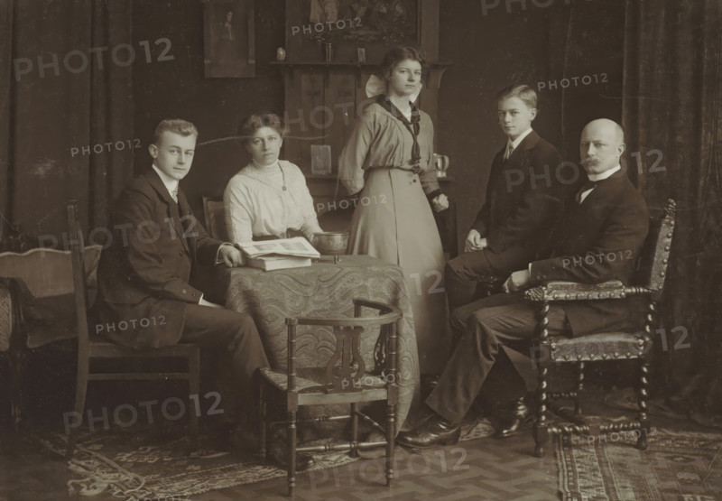 Portrait of a family in a homely environment, probably related to the Marmelstein family, photograph, inscription: 'Familie ...', photographer: Peter Clausing, Haarlem, 1895 - 1920, paper, pasteboard, baryta paper, matte daylight collodion silver print, height 173 mm x width 255 mm, historical persons, adult woman, adult man, A group portrait shows five people posed in a room with dark curtains and a patterned rug on a wooden floor. Three men and two women are present; three are seated and two are standing. The seated man on the left wears a dark suit and sits on a simple wooden chair beside a small table covered with a patterned cloth and stacked books or papers. A seated woman next to him wears a light-colored high-neck blouse and a dark belt, with a shallow bowl on the table beside her. A standing woman behind the table wears a light-colored skirt and a blouse with a dark scarf tied at the neck. A seated young man to the right wears a dark suit and sits on a carved wooden chair, whi...