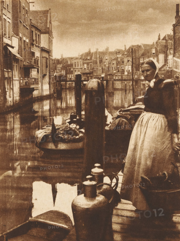 Woman with a basket on a pier in a harbor, photograph, 1850 - 1900, photographer, paper, pasteboard, salted paper print, height 234 mm x width 143 mm, adult woman, harbor, A sepia-toned scene of a canal lined with narrow, tall buildings along the left and distant background, their windows and balconies reflecting in the water; a docked boat filled with goods is moored near a wooden piling in the midground, and a woman in traditional clothing and a headscarf stands on the right near the water's edge with one arm resting on a post and a long skirt falling to her ankles; various jugs and containers sit on the wooden dock in the foreground, and additional small boats and rigging are visible along the canal with rooftops and chimneys forming the skyline under a cloudy sky.