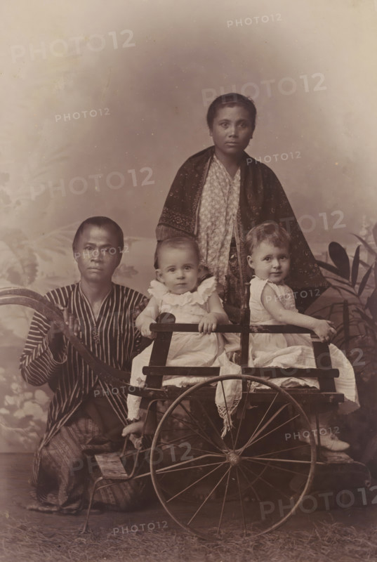 Portrait of two children, presumably the children of AF Marmelstein, with two servants, cabinet photograph, inscription: 'C. Nieuwenhuis Padang, Photographer. Sumatra's West Coast.', annotation: 'Hector & Hermien with, their nannies', number: '1899', photographer: Christiaan Benjamin Nieuwenhuis, Padang (city), 1891 - 1905, Christiaan Benjamin Nieuwenhuis, photographer, Sumatra, paper, pasteboard, daylight collodion silver print, height 165 mm x width 107 mm, historical persons, historical persons - BB - woman, child, A sepia-toned studio portrait shows four people arranged around a wooden cart with large spoked wheels; two small children in light-colored, frilly dresses sit side by side on the cart, each holding the cart's wooden rail, while an adult kneels to the left wearing a striped garment and resting a hand on the cart's handle, and another adult stands behind the children draped in a patterned shawl over a patterned dress.
