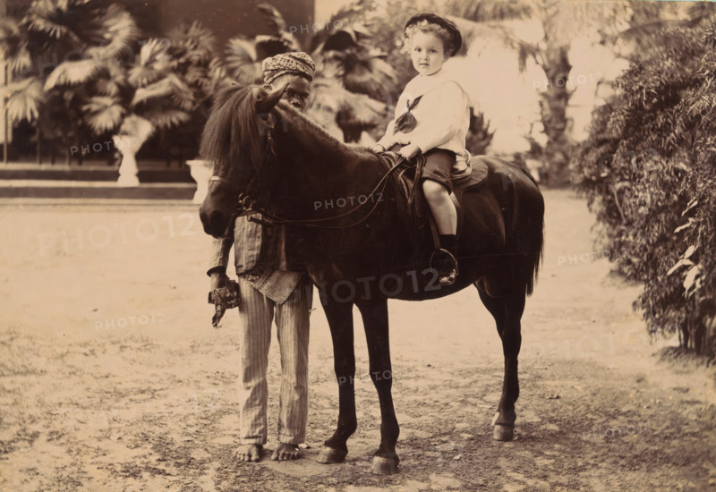 Portrait of an unknown child on a small horse with a servant, cabinet photograph, Illustration of a woman with a map in her hand and a landscape on the verso., photographer: Wah Seng, Makassar, 1905 - 1920, South Sulawesi, Sulawesi, paper, pasteboard, gelatin silver print, height 106 mm x width 166 mm, historical persons not known by name, horse, child between toddler and youth (of undetermined sex), A sepia-toned photograph shows a child seated on a dark horse, wearing a light-colored blouse with a tied scarf and a dark hat, with legs visible and feet in dark shoes resting in stirrups; the horse has a long mane partially covering its face and is fitted with a saddle and reins. Beside the horse stands a barefoot person holding the horse’s reins and wearing striped trousers and a patterned headwrap, with part of their upper body partially obscured by the horse. The background includes large leafy plants and palm fronds, a trimmed hedge on one side, and a flat ground surface in the foreg...
