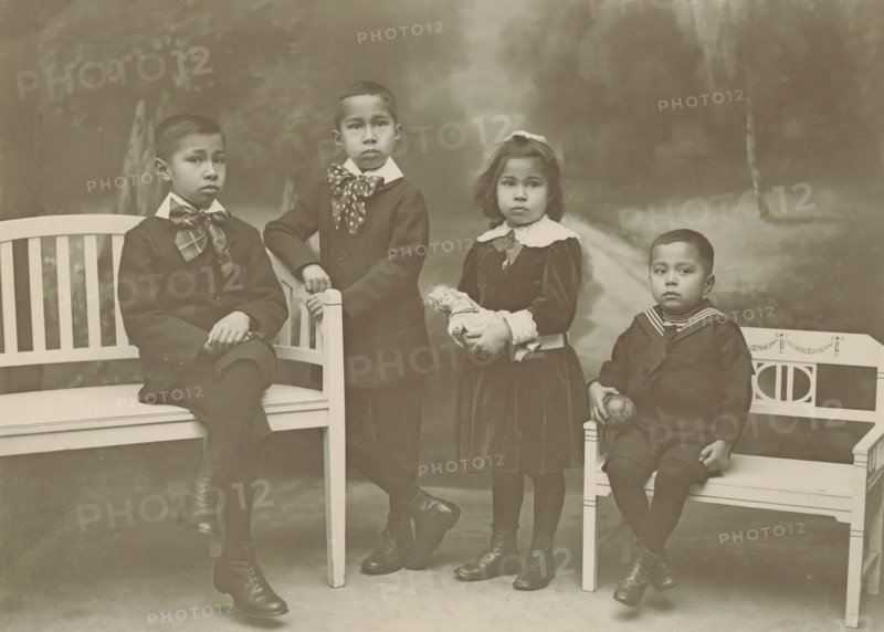 Portrait of four unknown children, probably related to the Marmelstein family, photograph, inscription: '..., Marmelstein-...', photographer: Friedrich Carel Hisgen, Amsterdam, photographer: Julius Heinrich Hisgen, 1888 - c. 1930, paper, pasteboard, baryta paper, gelatin silver print, developing gelatin silver print, height 240 mm x width 319 mm, historical persons, child, Four children are positioned on and between benches in a studio setting. On the left a boy sits with one leg crossed, wearing a dark suit and a large patterned necktie, his hands resting one over the other on his knee. Standing next to him is another boy dressed in a dark coat with a contrasting collar and a spotted scarf tied at the neck, one hand resting on the bench back and the other holding the bench post. Beside him a girl stands in a dark dress with a wide white collar and lace cuffs, holding a small doll in her hands and wearing a bow in her hair. On the right a younger boy sits on a smaller bench wearing a s...