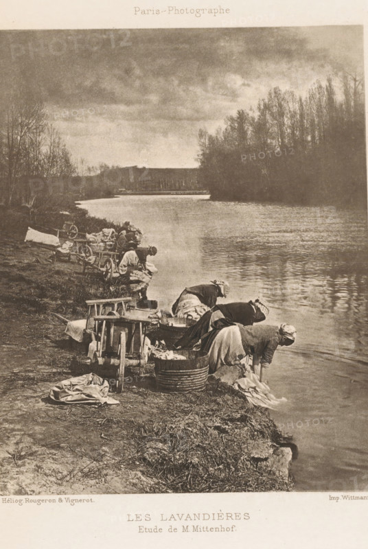 Les lavandières, Women doing laundry on the banks of a river, page, photomechanical print, photographer: M. Mittenhof, France, cliché maker: Rougeron, Vignerot et Cie, printer (person): Chardon-Wittmann (possibly), c. 1883 - in or before 1893, printer (person), paper, photoaquatint, height 150 mm x width 120 mm, river, laundering, A sepia-toned scene shows a riverbank with several women kneeling and bending over near the water, washing textiles in wooden tubs and basins; some garments and cloths lie on the ground and are draped over small wooden wringers and carts nearby. The river occupies the right side of the image, its surface reflecting the overcast sky. Bare trees and dense foliage line the far bank, extending toward the horizon, while the near bank is grassy and uneven with scattered laundry tools and wicker baskets. The overall atmosphere is quiet and focused on the washing activity along the shoreline.