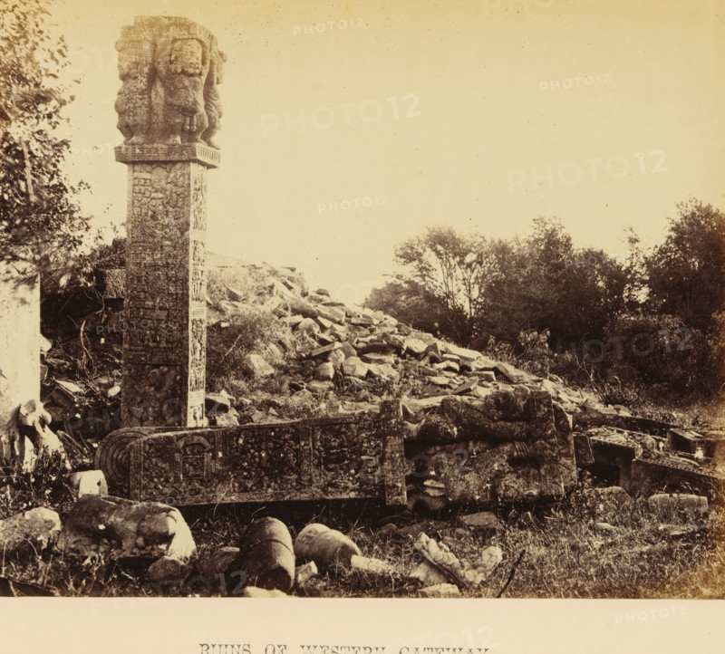 Ruins of the western gateway (torana) of the Great Stupa at Sanchi, Madhya Pradesh, India, photograph, page, inscription: 'SANCHI. PLATE XIX., RUINS OF WESTERN GATEWAY.', photographer: James John Waterhouse, Sanchi, c. 1863 - in or before 1868, Central India, paper, albumen print, height 166 mm x width 213 mm, Photo two pages after page 108., gate, entrance, metropolis, places and objects of worship, Hinduism, Buddhism, Jainism, A tall carved stone pillar stands on the left with sculpted figures near its top and relief patterns along its shaft, adjacent to a low carved stone wall or barricade with ornate panels and circular motifs; in front of these structures lie scattered broken stone fragments and rounded cylindrical pieces on grassy ground, and behind them a slope covered in rubble and rocks rises toward a cluster of trees and shrubbery against a pale sky.