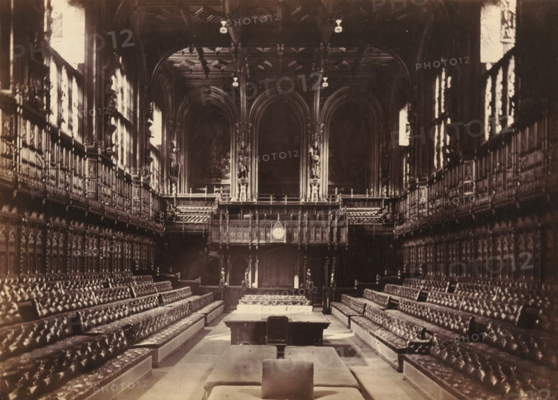 View of the interior of the House of Representatives, photograph, page, photographer: John Harrington, London, c. 1864 - in or before 1869, paper, albumen print, height 140 mm x width 194 mm, palace, interior, representation of a building, Houses of Parliament, A long, ornate hall viewed from one end toward a raised, carved wooden screen and balcony with tall arched openings and decorative columns, flanked on both sides by high, paneled walls with rows of tufted leather benches facing inward, patterned upholstery visible on the seating, a central aisle leading to a table and padded benches in the foreground, tall windows along the side walls admitting bright light, intricate woodwork and sculpted figures incorporated into the balcony and wall panels, and a clock set into the central wooden screen above the entrance.