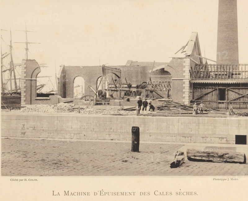 View of a machine in the remains of a munitions factory that exploded in Oosterweel near Antwerp in 1889, La machine d'épuisement des cales sèches, page, photomechanical print, number: '6', photographer: Henri Colon, Oosterweel, cliché maker: anonymous, Belgium, printer (person): J. Maes (possibly), Antwerp, publisher: J. Maes, 1889, Henri Colon, J. Maes, photographer, cliché maker, printer (person), publisher, paper, collotype, light printing, height 165 mm x width 230 mm, Image on page 6., ruin of a building, architecture, machines, parts of machines, tools and appliances, Explosion of a munitions factory in Oosterweel in 1889, A wide sepia-toned scene of a waterfront construction area shows a partially built or demolished brick structure with large arched openings and exposed wooden beams, adjacent to a tall cylindrical chimney. Scattered piles of rubble and stacks of timber lie along the quay in front of the building, where scaffolding, ladders, and construction machinery with visi...
