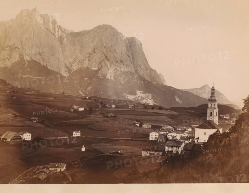 View of Mount Schlern in the Dolomites, Italy, Le Schlern, photograph, page, number: 'N. 14', photographer: Eugène Goblet d'Alviella (possibly), Dolomites, c. 1873 - in or before 1878, Eugène Goblet d'Alviella, paper, albumen print, height 186 mm x width 265 mm, prospect of village, silhouette of village, mountains, Schlern, A sepia-toned landscape showing a village in a valley with rolling fields and scattered farmhouses, a tall church tower with a rounded multi-tiered spire near the right edge, clusters of houses with pitched roofs in the foreground, terraced fields and small isolated buildings across the midground, and a sheer rocky mountain face rising behind the valley and occupying much of the left and central background under a pale sky.