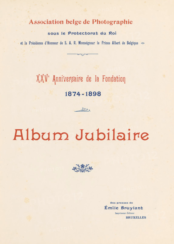 XXVe anniversaire de la fondation, 1874-1898 : album jubilaire, book, date: 'July 1985', number: '1000', 1898, textile materials, paper, linen (material), transparent paper, printing, autotype, height 288 mm x width 227mm x thickness 18mm, A cream-colored page with small scattered brown spots and a large decorative heading in a reddish-brown serif type near the top, a smaller line of dark blue ornate type above it and another line of dark blue type centered below, a small blue decorative flourish between lines, a larger reddish-brown title in the middle of the page, a small blue floral ornament beneath it, and a block of small dark blue type near the lower right portion of the page.