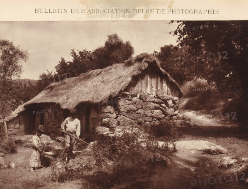 Unknown man and girl at a house, page, photomechanical print, inscription: 'Bulletin de l'association belge de photographie, Concours de négatifs 1888. (Médaille d'Argent)', photographer: Joaquim Basto, cliché maker: anonymous, c. 1883 - in or before 1888, Joaquim Basto, photographer, cliché maker, paper, Woodburytype, height 115 mm x width 166 mm, Print two pages for page 622., historical persons, housing, A sepia photograph showing a rustic stone cottage with a thatched roof set among trees and bushes, a dirt path winding past the cottage, a man standing near the cottage holding a long tool, a young girl standing nearby holding a basket, visible stone walls and a wooden gable, leafy branches framing the scene, and textured ground and vegetation in the foreground.