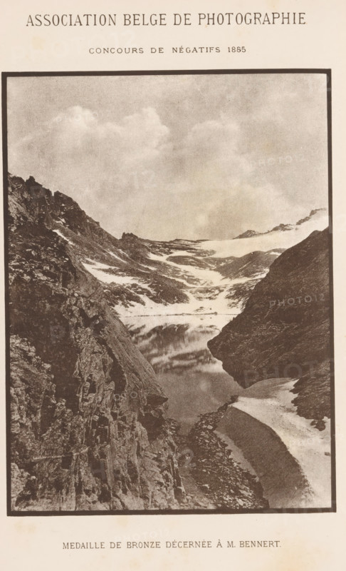 View of a water in the mountains, page, photomechanical print, maker: Angerer & Göschl, C., c. 1880 - in or before 1885, paper, autotypie, height 163 mm x width 122 mm, Print two pages before page 436., landscapes with waters, waterscapes, seascapes (in the temperate zone), mountain-top, peak, A vertically oriented sepia-toned photograph showing a steep rocky mountain valley with jagged cliffs on either side, patches of snow and ice clinging to slopes, a narrow reflective body of water nestled between the rocks, and a sky filled with soft clouds above.