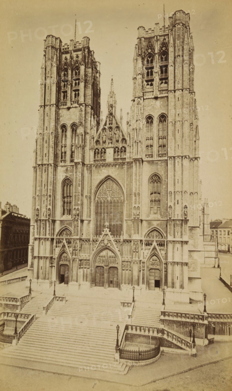Bruxelles. Façade de Ste-Gudule, Cathedral of Saint Michael and Saint Gudula in Brussels, photograph, page, Brussels, c. 1875 - c. 1880, photographer, paper, albumen print, albumendruk, height 172 mm x width 102 mm, church (exterior), facade (of house or building), Cathedral of Saint Michael and Saint Gudula, A sepia-toned photograph of a large Gothic cathedral façade with two tall towers flanking a central pointed arch window and ornate sculpted gables, detailed tracery and vertical buttresses. The lower section shows three large pointed doorways with carved tympana and decorative niches above, multiple narrow lancet windows higher up, and rows of sculpted figures in recessed panels. Broad stone steps lead up to the entrance, bordered by balustrades and lamp posts, with smaller staircases and fenced curved landings at the sides. Adjacent buildings and a street are visible at the edges of the image.