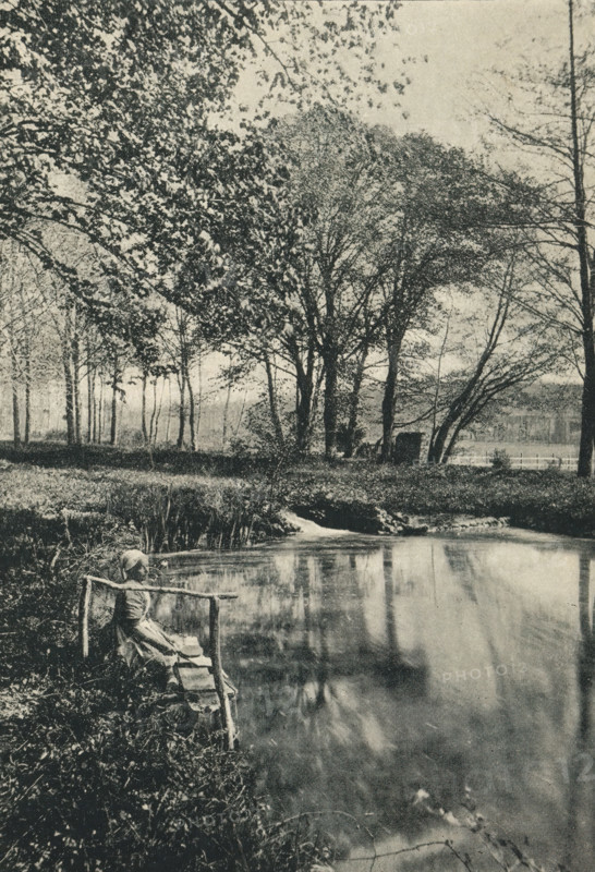 L'Étang de Bel-Air, Près Tours, Woman on the bank of a water feature, page, photomechanical print, inscription: 'Papier &lt, Lustré &gt, . Grosvenor, Chater et Co, 6, rue Favart, Paris.', photographer: Rougé, Tours, cliché maker: Ducourtioux & Huillard, c. 1891 - in or before 1896, Rougé, Ducourtioux & Huillard, photographer, cliché maker, paper, autotype, zincogravure, height 160 mm x width 110 mm, water course (landscape with figures, staffage), A grainy black-and-white scene of a riverside with tall trees along the far bank, their branches and leaves forming a dense canopy that extends across the top of the image, and a calm stretch of water in the foreground reflecting the trees; a small rustic wooden railing and a narrow bench sit at the near edge of the water, where a seated figure in plain clothing and a head covering rests with arms extended to the railing; grasses and low vegetation grow along the riverbank and a small patch of reeds projects into the water, while trunks and t...