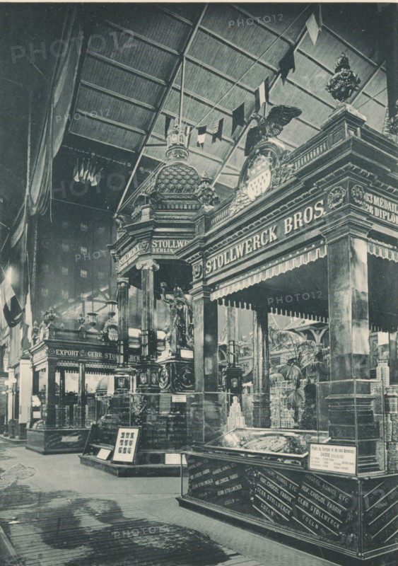 Pavillons en Chocolat (Gebr. Stollwerck, Koeln), Pavilions of the Stollwerck brothers during the 1894 World's Fair in Antwerp, photomechanical print, Part of a cover with 49 plates., inscription: 'Exposition Universelle D'Anvers 1894', photographer: Charles Bernhoeft, Antwerp, Luxembourg (city), c. 1894, printer (person), paper, collotype, photogravure, height 215 mm x width 153 mm, stall, booth (, market), world fair, world exhibition, Wereldtentoonstelling 1894 (Antwerp), Period, 1894, A large indoor scene shows an ornate exhibition space with tall decorative columns, sculpted figures, and elaborate architectural details topped by domes and cresting. A high arched roof with visible rafters spans overhead, and flags or banners hang from above. Carved statues and a winged figure appear atop pedestals, and display cases with angled glass tops contain objects. Potted plants and smaller ornamental elements fill the area, and a patterned wooden floor runs through the foreground. A grand po...