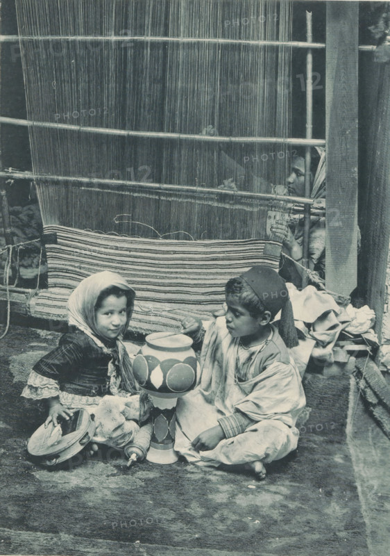 Two unknown Tunisian children in front of an unknown woman weaving a carpet, Tunisia - fabricante de tapis, photomechanical print, Part of a cover with 49 plates., inscription: 'Exposition Universelle D'Anvers 1894', photographer: Charles Bernhoeft, Antwerp, cliché maker: Charles Bernhoeft, Luxembourg (city), c. 1894, Charles Bernhoeft, photographer, cliché maker, printer (person), paper, collotype, photoprint, height 215 mm x width 153 mm, child (at home), Asiatic races and peoples (with NAME), tapestry weaver, gobelin maker, world fair, world exhibition, World Exhibition 1894 (Antwerp), Period, 1894, A woven vertical loom fills the upper background with thin vertical threads and horizontal wooden rods, and a partially completed striped textile hangs from it; in front of the loom two children sit on the ground holding a painted tall ceramic object, the child on the left wearing a headscarf and layered clothing with lace cuffs and holding a small tambourine-like instrument, the child o...