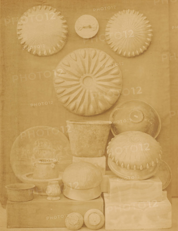 Silver dishes and bowls in the Boulaq Museum in Cairo, Egypt, photograph, page, photographer: Hippolyte Délié, Cairo (city), photographer: Emile Béchard, c. 1866 - in or before 1871, Ottoman Empire, North Africa, paper, albumen print, height 240 mm x width 183 mm, permanent exhibition, museum, bowl, dish, plate, saucer, Boulaq Museum, A vertical arrangement of sculpted round and bowl-shaped objects in a monochrome sepia tone fills the frame, including several large circular discs with radial petal-like patterns, a smaller disc with a handle-like raised feature, domed forms with scalloped edges, shallow bowls and pots, a tapered cylindrical vessel, and stacked rectangular blocks used as supports, all showing textured surfaces and subtle shadows against a plain background.