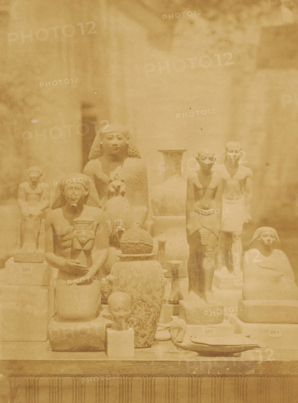 Images from various eras and styles in the Boulaq Museum in Cairo, Egypt, photograph, page, photographer: Hippolyte Délié, Cairo (city), photographer: Emile Béchard, c. 1866 - in or before 1871, Ottoman Empire, North Africa, paper, albumen print, height 232 mm x width 174 mm, piece of sculpture, reproduction of a piece of sculpture, permanent exhibition, museum, Boulaq Museum, A sepia-toned photograph shows a display of carved stone figures and artifacts arranged on tiered platforms, with several human-shaped statues in various seated and standing poses, some wearing headdresses and one holding a small figure; there are rounded vessels, a woven or textured spherical object, and other sculpted items placed among the figures, all set against a plain background with soft shadows.