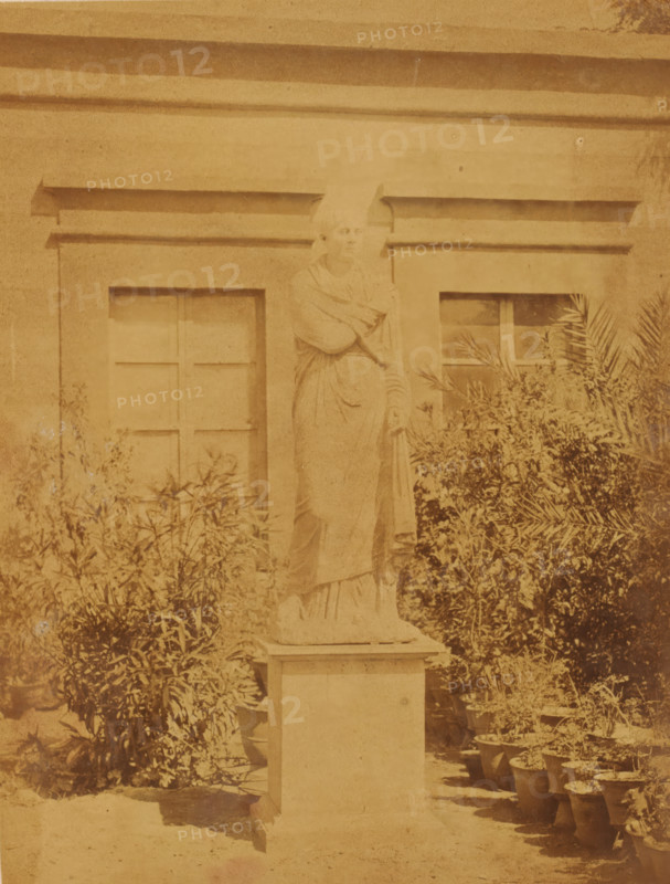 Roman marble statue, found in Tell Mokdam, in the garden of the Boulaq Museum in Cairo, Egypt, photograph, page, photographer: Hippolyte Délié, Cairo (city), photographer: Emile Béchard, c. 1866 - in or before 1871, Ottoman Empire, North Africa, paper, albumen print, height 241 mm x width 184 mm, permanent exhibition, museum, piece of sculpture, reproduction of a piece of sculpture (Roman art), Boulaq Museum, A weathered stone statue of a robed figure stands on a rectangular pedestal in front of a building facade with two recessed window frames; the figure's head is slightly tilted and one arm is raised across the chest while the other rests downward holding a draped garment. Shrubbery and potted plants surround the base of the pedestal and line the ground to the right, with foliage partially obscuring the lower windows. The image has an overall sepia tone and shows signs of age with uneven shading and surface marks across the scene.