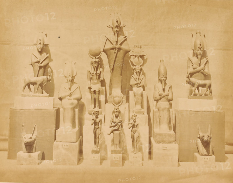 Statues of Osiris and other Egyptian gods in the Boulaq Museum in Cairo, Egypt, Statues of Osiris and other Egyptian idols in the Boulaq Museum in Cairo, photograph, page, photographer: Hippolyte Délié, Cairo (city), photographer: Emile Béchard, c. 1866 - in or before 1871, Ottoman Empire, North Africa, paper, albumen print, height 193 mm x width 252 mm, permanent exhibition, museum, Osiris, god of the underworld, statues, paintings, etc., objects of worship (Egyptian religion), Boulaq Museum, A row of carved figures and animal sculptures arranged on tiered rectangular plinths against a plain wall, each figure carved in a stylized, upright pose with some seated and some standing, many wearing tall headdresses and holding staffs or crossed arms, and two animal sculptures resembling reclining or seated jackal-like heads positioned at the lower corners.