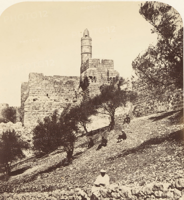 View of the Tower of David in Jerusalem, From the SW outside the Wall of the City, Citadel, photograph, page, photographer: James McDonald, Jerusalem, c. 1860 - in or before 1865, paper, albumen print, height 523 mm x width 340 mm, Photo on page 19., fortified city, Tower of David, A sepia-toned photograph showing a stone fortress wall with a cylindrical tower rising above a crenellated section, framed by trees with dark foliage on the right and scattered trees on a sloping rocky hillside in the foreground, where several seated figures in dark clothing are positioned among the stones and a single figure in lighter clothing sits closer to the bottom edge.