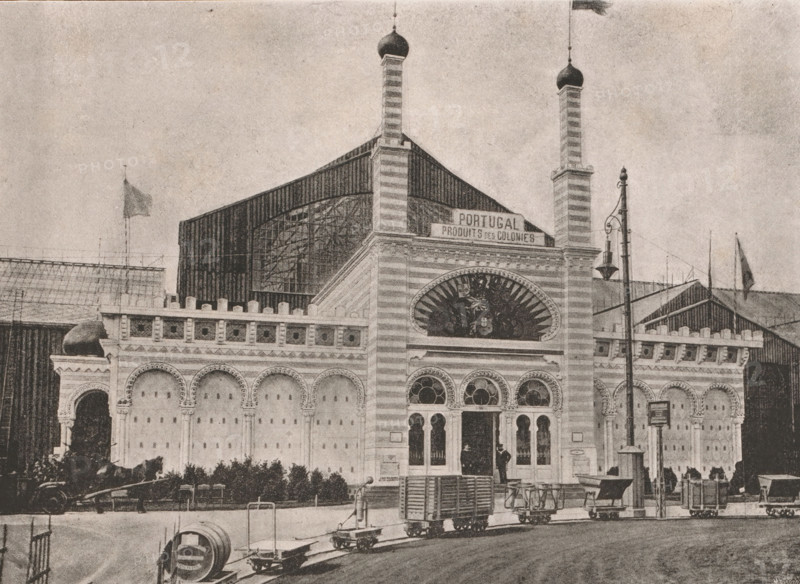 View of the pavilion of the Portuguese colonies at the 1885 World's Fair in Antwerp, Façade du Pavillon des Colonies Portugaises, page, photomechanical print, Antwerp, clichémaker: Meisenbach's Autotype Company, c. 1880 - in or before 1885, Meisenbach's Autotype Company, photographer, clichémaker, paper, autotypie, height 167 mm x width 233 mm, world fair, world exhibition, Wereldtentoonstelling 1885 (Antwerp), Period, 1885-05-02 - 1885-11-02, A large ornate building facade with striped vertical towers topped by bulbous domes, arched windows and decorative circular windows, a semicircular decorative panel above the entrance, a row of arched recesses along the wall, and columns with ornate capitals; a tall lamp post with a hanging light stands near the entrance; several small wheeled carts and platforms line the street in front, along with a cylindrical object on the pavement; flags fly on poles above parts of the structure and nearby roofs; a person stands in the entranceway and anothe...