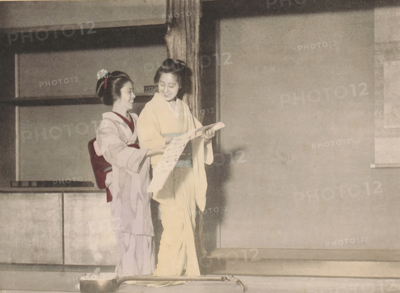 Two geishas with a letter, The Geishas Holding a Letter, page, photomechanical print, Japan, cliché maker: Kazumasa Ogawa, Tokyo, c. 1895 - in or before c. 1900, photographer, Kanto, paper, collotype, color lithography, hand coloring, key in light print with color lithography and partly hand colored, height 167 mm x width 230 mm, courtesan, hetaera, adult woman (two persons), Two women wearing traditional robes stand close together near a wooden post; one woman wears a light-colored robe with a pale sash and holds a long narrow scroll while the other woman, in a patterned robe with a darker sash and a hair ornament, stands slightly behind her smiling and looking at the scroll; the scene shows a simple interior with plain walls, sliding panels, and a low platform, and a small lacquered box with a handle rests on the floor in front of them.