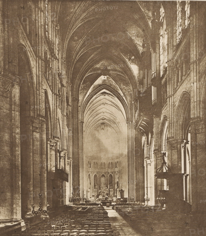 Chatres Cathedral, high altar, Interior of Chartres Cathedral, page, photomechanical print, cliché maker: Lakeside Press, publisher: Haskell & Post Publishers, c. 1881 - in or before 1886, photographer, paper, collotype, photoprint, height 229 mm x width 204 mm, interior of church, altar, Chartres, Chartres Cathedral, A sepia-toned interior view of a large cathedral nave with tall pointed vaulted ceilings and a central aisle flanked by rows of wooden pews, high clustered columns and rounded arches lining both sides, ornate stonework and carved details on the pillars, large stained glass windows set into the upper walls on the right, a raised altar area at the far end with sculptural elements and ornamental screens, hanging chandeliers and metal candle stands visible near the front and along the sides, and a pulpit or raised lectern projecting from the right supported by decorative brackets.