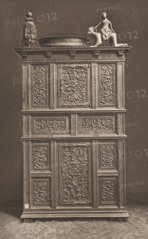 15th century German wooden cabinet, at the 1877 Exposition Rétrospective in Lyon, France, page, photomechanical print, inscription: 'ARMOIRE, ALLEMAGNE_FIN DU XVe SIÈCLE, COLLECTION DE Mr CHABRIÈRES-ARLES.', inscription: 'EXPOSITION RÉTROSPECTIVE DE LYON, PAR JB GIRAUD.', number: 'PL. I.', Lyon, cliché maker: Dujardin, printer (person): Imprimerie Eudes, Paris, 1877 - in or before 1880, photographer, printer (person), paper, photoaquatint, height 233 mm x width 147 mm, ornamental cupboard (wood and other phytogenic material), A tall wooden cabinet with multiple carved panels occupies the frame, topped by a decorative arrangement that includes an ornate metal lantern, a shallow round basin with embossed patterns, and a sculptural figure of a person seated on a stylized animal; the cabinet surface shows many rectangular framed panels filled with intricate scrolling foliage carvings and decorative moldings, metal hinges and latches are visible on some panels, the wood has a worn, aged app...