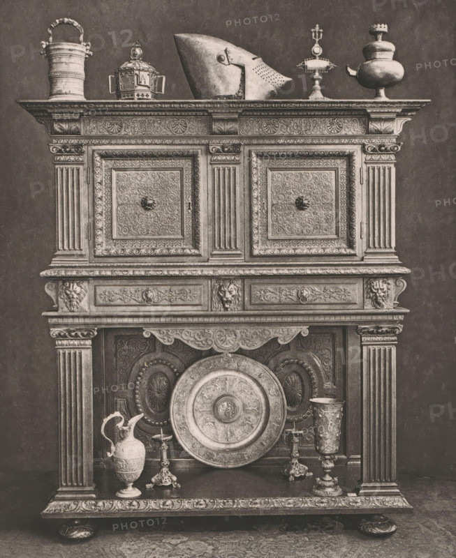 16th century French wooden sideboard, at the 1877 Exposition Rétrospective in Lyon, France, page, photomechanical print, inscription: 'CRÉDENCE, FRANCE_XVIe SIÈCLE, COLLECTION DE Mr CHABRIÈRES-ARLÈS.', number: 'PL. XXII.', inscription: 'EXPOSITION RÉTROSPECTIVE DE LYON, PAR JB GIRAUD.', Lyon, cliché maker: Dujardin, printer (person): Imprimerie Eudes, Paris, 1877 - in or before 1880, photographer, printer (person), paper, photoaquatint, height 227 mm x width 182 mm, ornamental cupboard (wood and other phytogenic material), A tall ornate wooden cabinet with carved panels and fluted columns displays a variety of metal objects on its top and lower shelf; the top surface holds a handled tankard, a domed container with side handles, a pointed helmet, a pedestal with a finial, and a bulbous vessel with a spout, all rendered in a tarnished metallic tone, while the lower shelf features a large decorative circular platter leaning against the back, a lidded ewer with a curved handle, two candles...