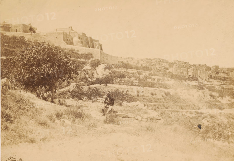 Bethlehem, with the church of nativity, Unknown man in a landscape with the Church of the Nativity in Bethlehem in the background, page, This photo is part of a cover with 7 sheets of albumen prints., photographer: Francis Frith, Jerusalem, 1857, paper, albumen print, height 158 mm x width 225 mm, Person, historical persons, church (exterior), city-view, and landscape with man-made constructions, Palestine, A wide sepia-toned landscape showing a hillside with stone terraces and scattered vegetation leading up to large stone buildings and walls on the left and a cluster of stone houses further back on the right, with a large leafy tree on the left midground and a person wearing layered clothing and a head covering seated on a low stone ledge near the center foreground.