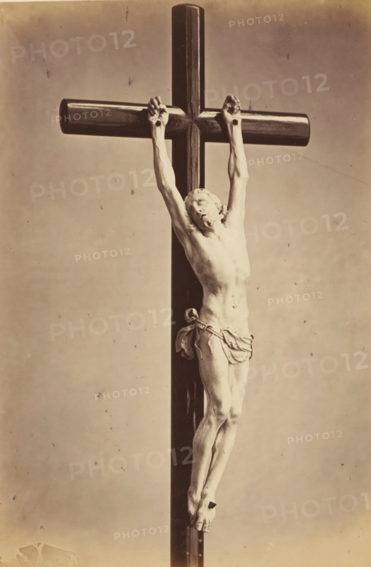Ivory crucifix from the episcopal chapel in Ghent, displayed at an exhibition of religious objects from the Middle Ages and Renaissance in 1864 in Mechelen, photograph, page, number: '8', photographer: Joseph Maes, Mechelen, 1864, paper, albumen print, height 277 mm x width 197 mm, altarcross, crucifix, piece of sculpture, reproduction of a piece of sculpture (bone, ivory, arts), Period, A sepia-toned photograph of a carved figure of a man affixed to a tall wooden cross, his arms extended upward and hands gripping the horizontal beam, his head tilted to one side with closed eyes, a cloth draped around his waist, detailed musculature and ribs visible on his torso, legs crossed at the ankles and feet overlapping where they are bound, the vertical post of the cross running from top to bottom behind him, the horizontal beam rounded and polished, and a plain neutral background with small specks and age marks.